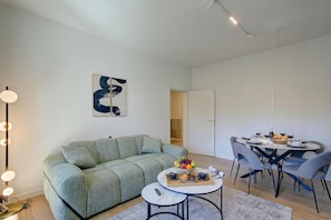 A modern living room with a green sofa, a round dining table with chairs, and a coffee table with a fruit bowl and cups. - Boutique Suites Two Bedroom Apartment Deluxe - AC (Lugano)