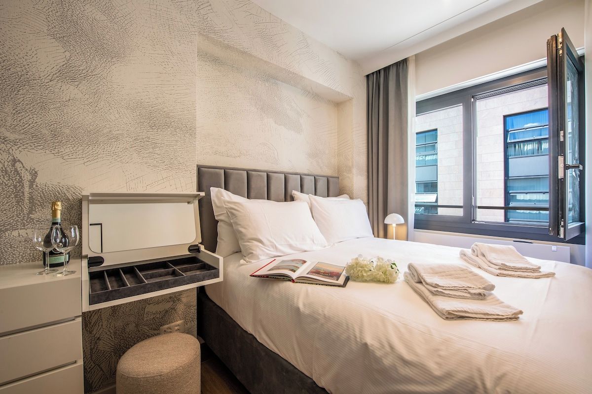 Une chambre d’hôtel avec un lit, une table de chevet sur laquelle se trouvent une bouteille et des verres, un livre et une fenêtre avec des rideaux.