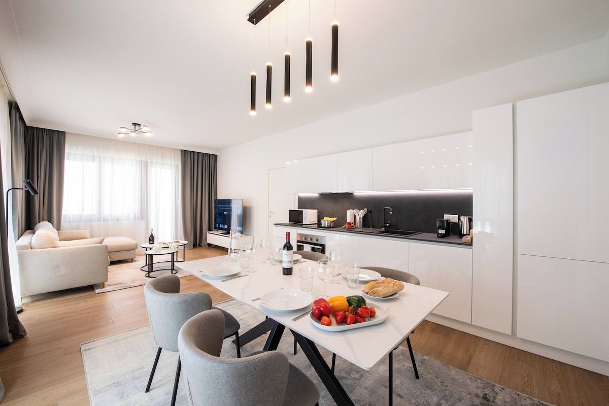 Ein modernes Wohnzimmer mit Sofa, Esstisch und Küchenzeile mit weißen Schränken sowie einem Flachbildfernseher.