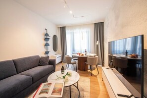 A modern living room with a sofa, a coffee table, a dining area with chairs, a flat-screen TV, and a large window with curtains. - Boutique Suites - Family Suite with AC (Lugano)