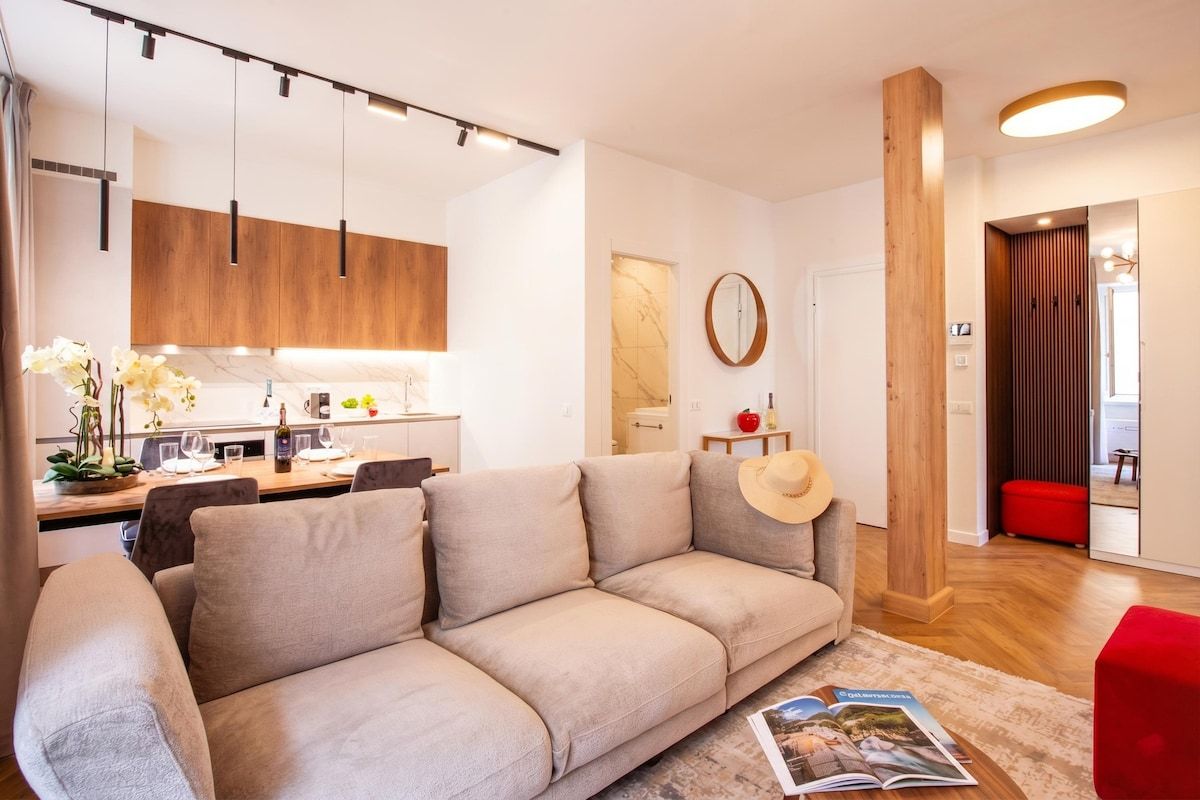 Una sala de estar moderna con un sofá color beige, una pared de acento de madera y una zona de cocina al fondo.