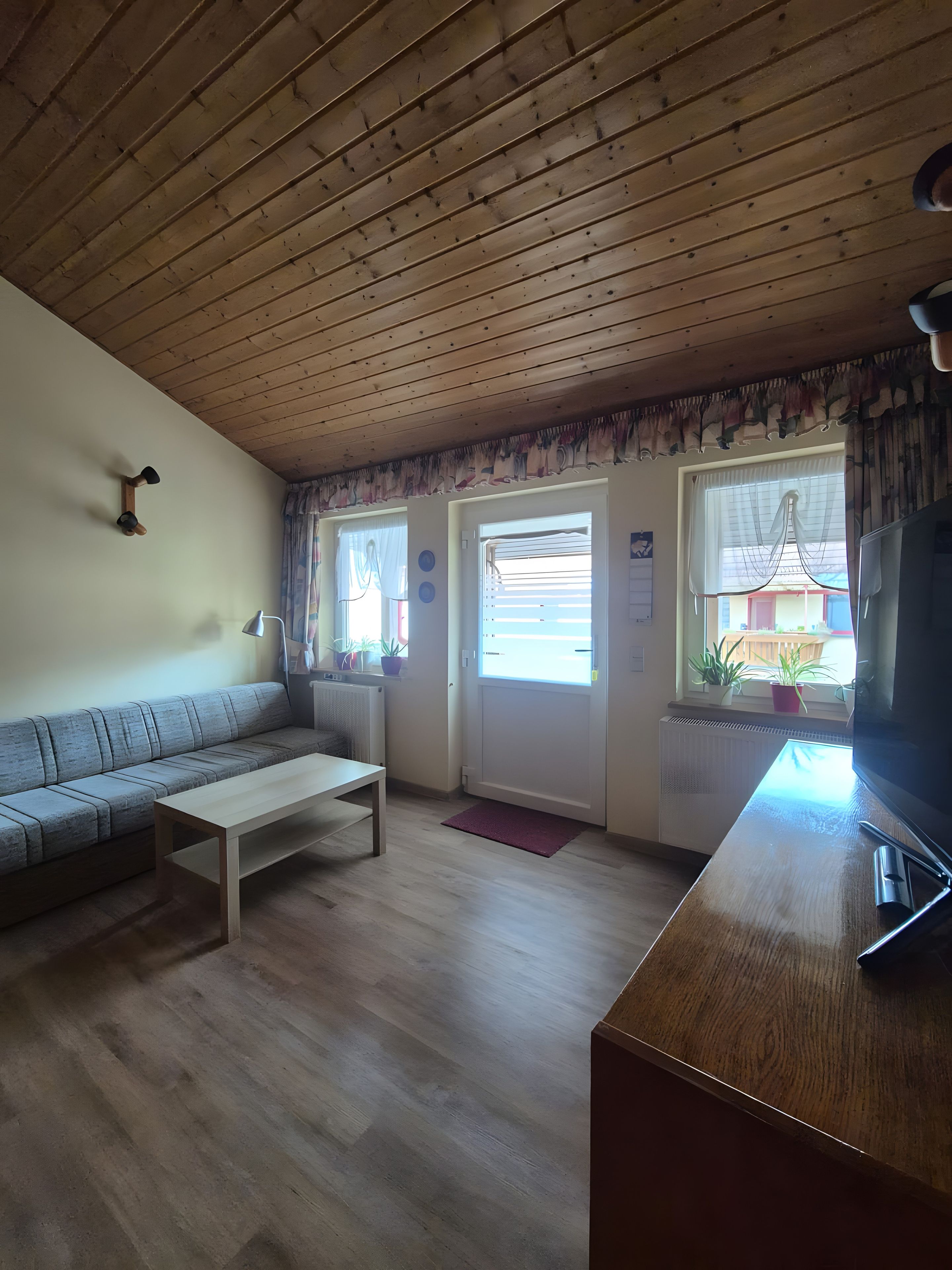 A living room with a wooden ceiling, a sofa, a coffee table, a television, and a window with curtains.