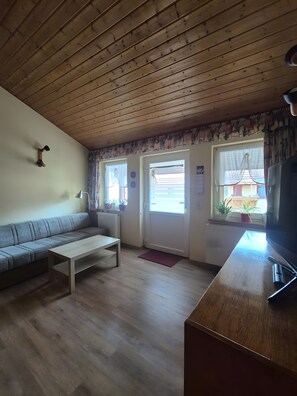 A living room with a wooden ceiling, a sofa, a coffee table, a television, and a window with curtains. - Small, Cozy Apartment with Country Charm, Shared Garden & Wi-Fi (FeWo 4) (Hörselberg-Hainich)