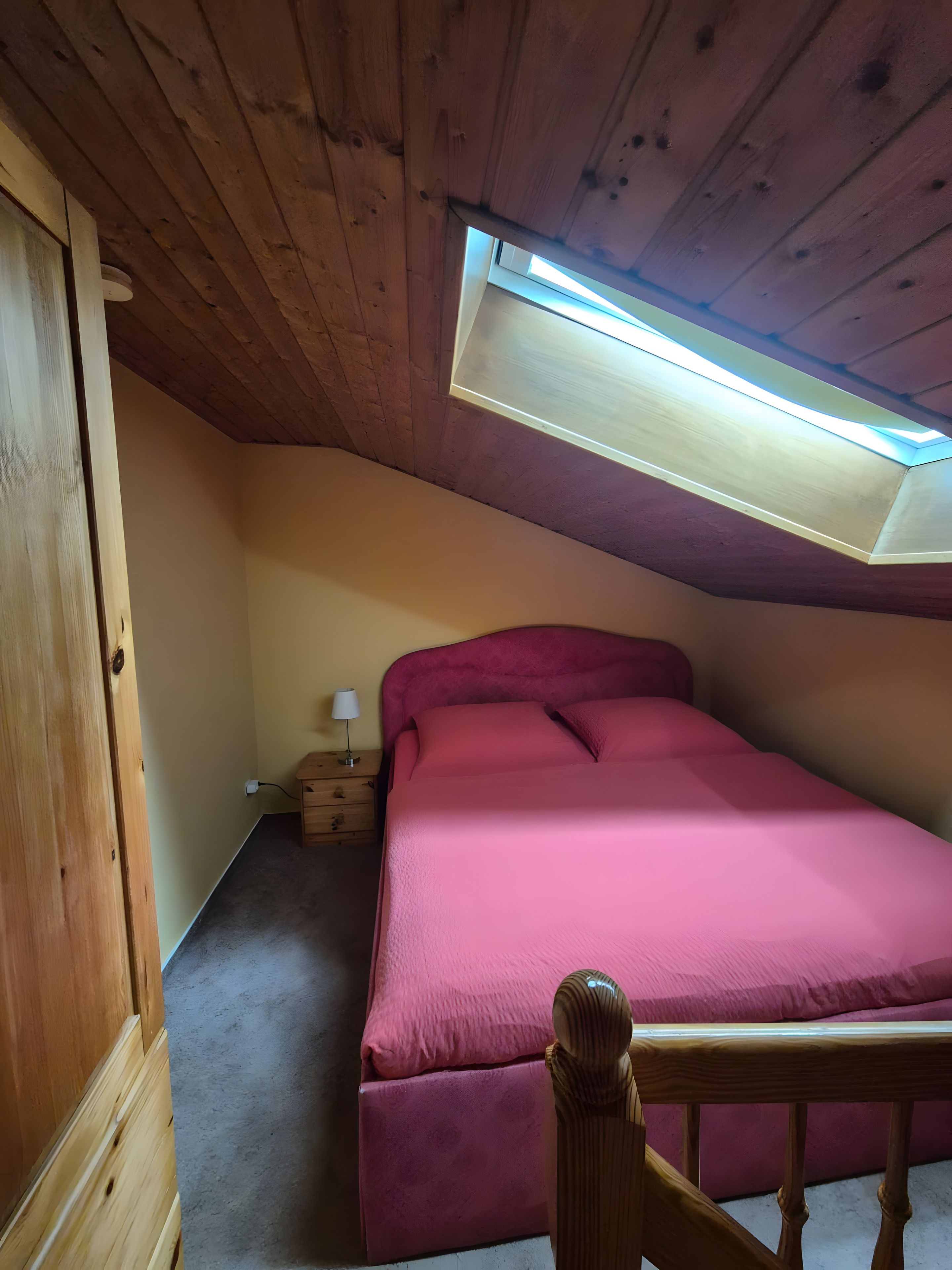 A bedroom with a sloped ceiling, a skylight, a bed with a pink bedspread, a nightstand with a lamp, and a wooden chair.