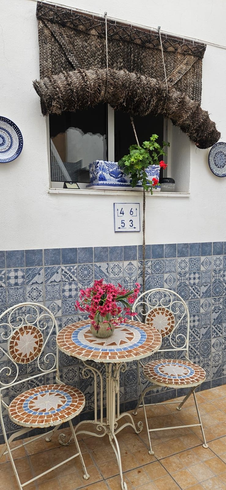 Un patio con una mesa y sillas, una maceta colgante con flores y una ventana con un toldo tejido.