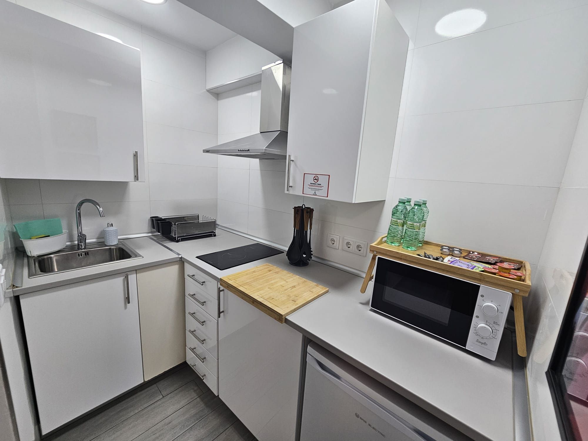 Cocina compacta con armarios blancos, un lavamanos, un microondas y una tabla de cortar de madera.