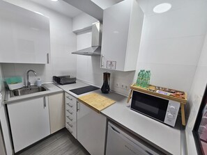 Cocina compacta con armarios blancos, un lavamanos, un microondas y una tabla de cortar de madera.