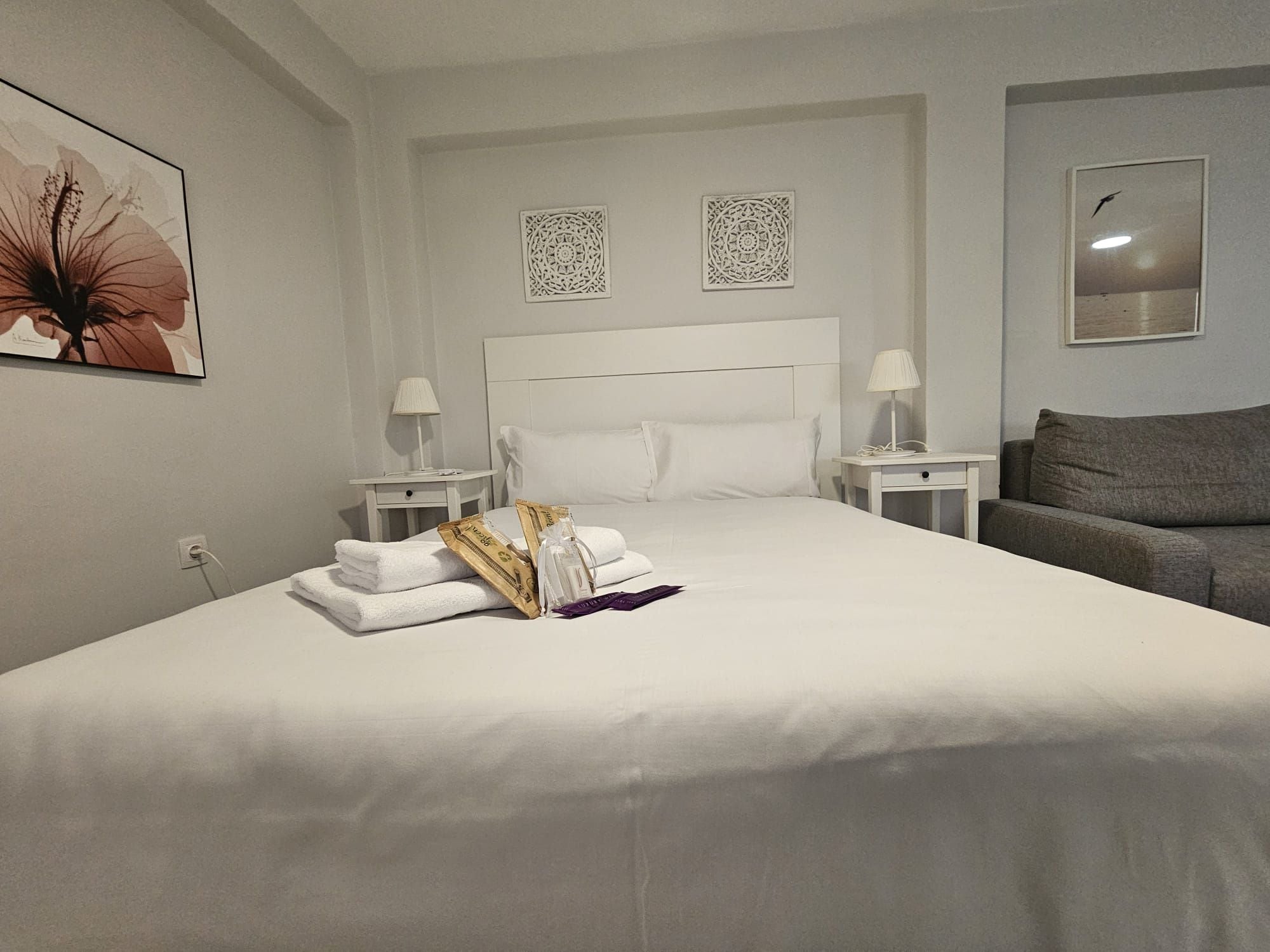 Une chambre d’hôtel avec un grand lit, deux tables de chevet avec des lampes, un canapé gris et des œuvres d’art décoratives au mur.