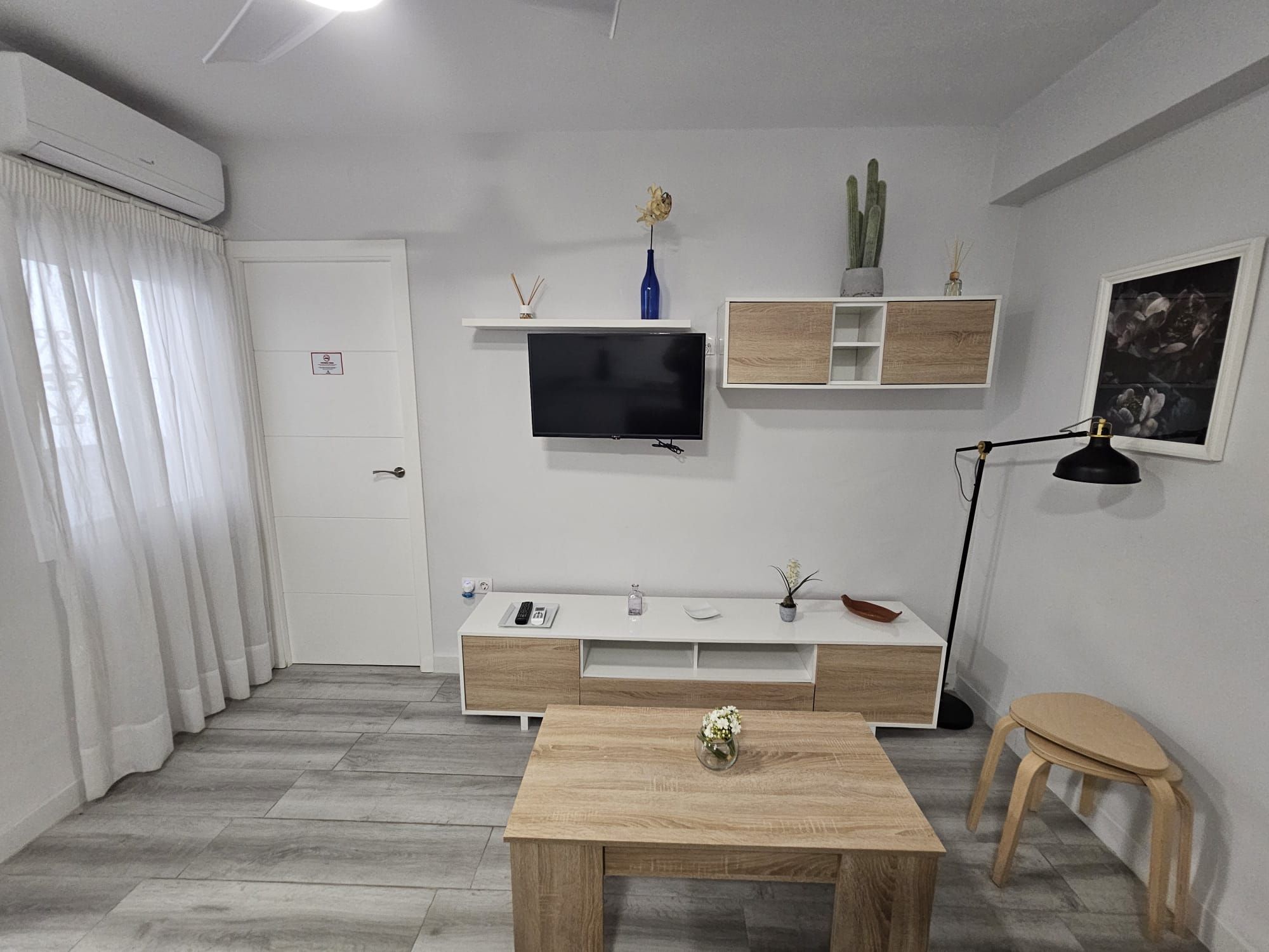 Een moderne woonkamer met een flatscreen televisie aan de muur, een houten salontafel, een klein bijzettafeltje, een vloerlamp en een minimalistische inrichting.