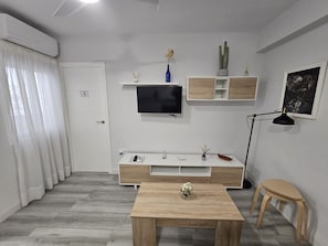 Una sala de estar moderna con un televisor de pantalla plana montado en la pared, una mesa de centro de madera, una pequeña mesita auxiliar, una lámpara de pie y un estilo decorativo minimalista.