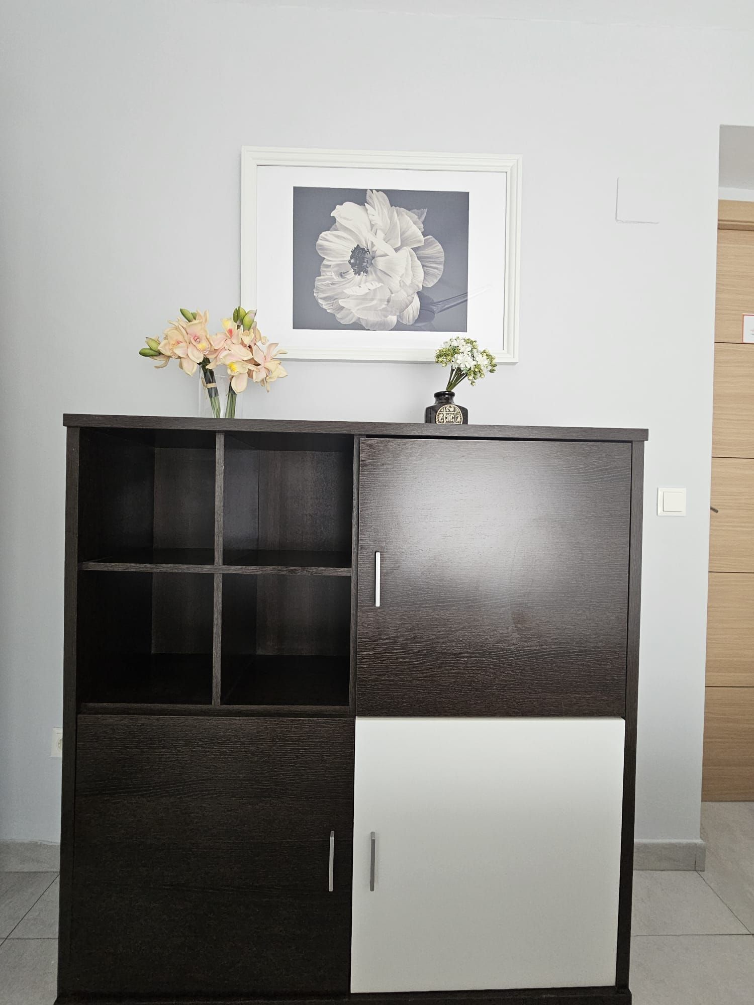 Un meuble en bois foncé, avec une partie inférieure blanche, deux tiroirs et une étagère. Un tableau floral encadré est accroché au-dessus du meuble.