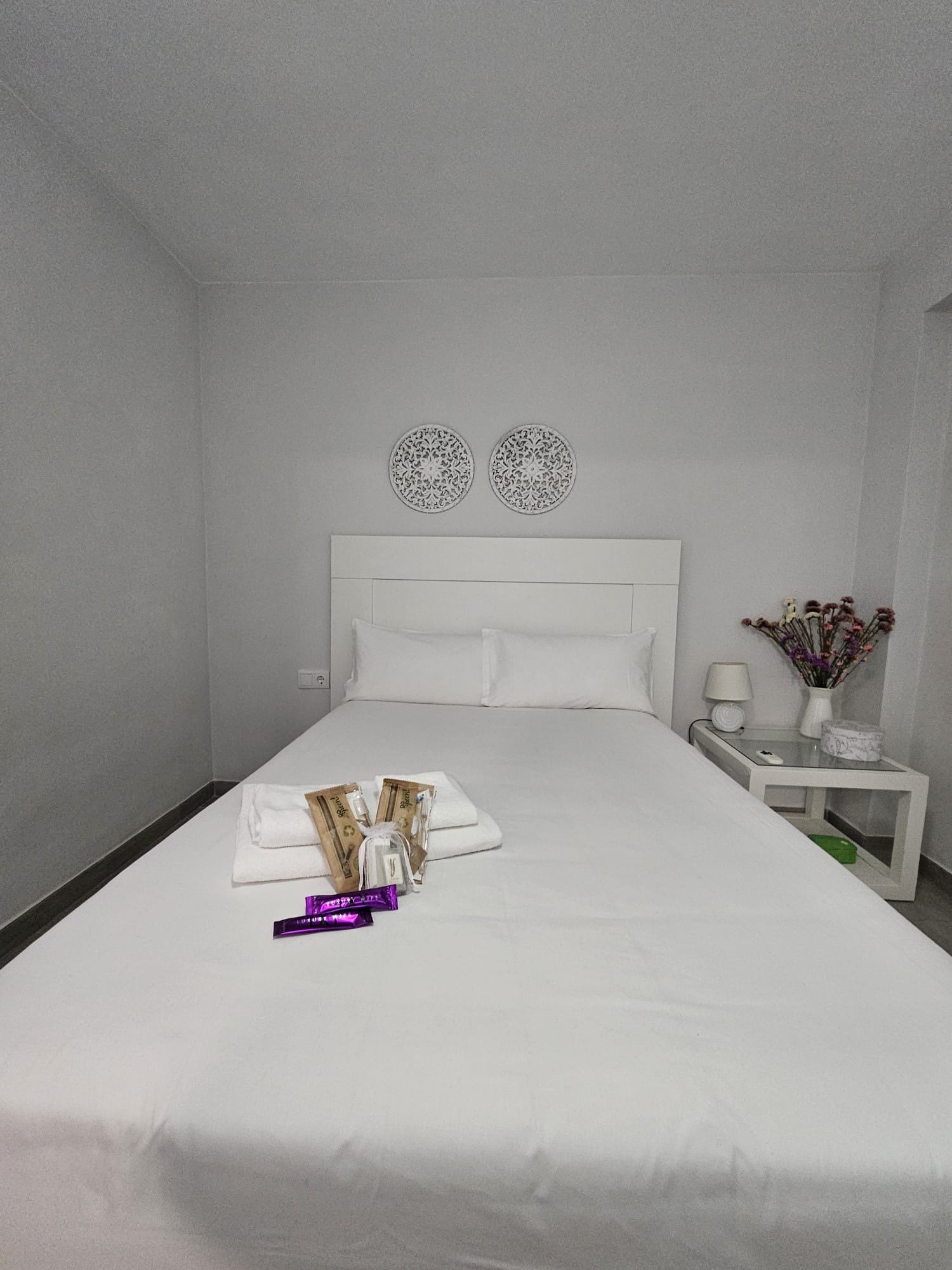 Una habitación de hotel con una cama grande, una mesita de noche con lámpara y un florero con flores.