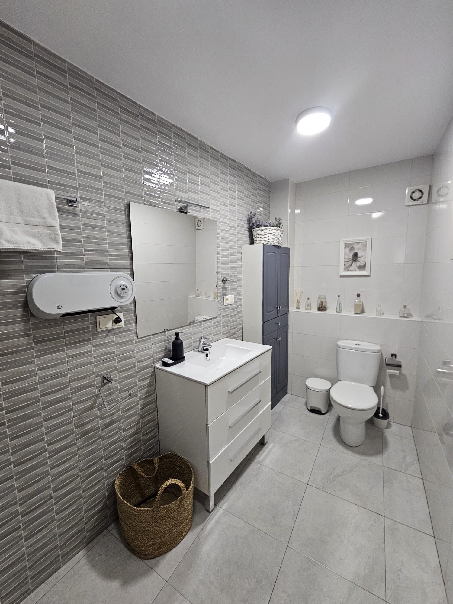 A modern bathroom with a white sink, mirror, and toilet. There is a basket on the floor and a small shelf with toiletries.
