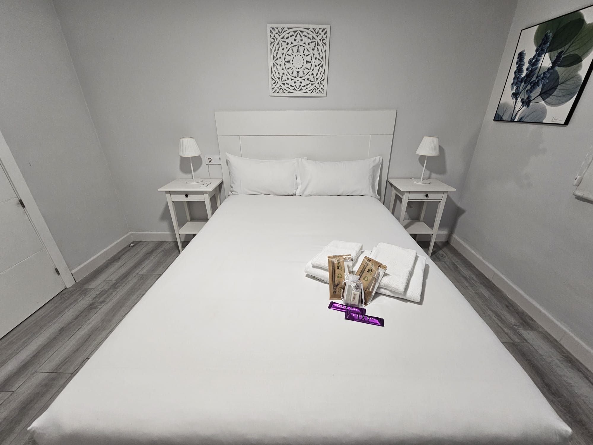 Una cama bien tendida con almohadas, dos mesitas de noche con lámparas y una obra de arte en la pared sobre la cama.