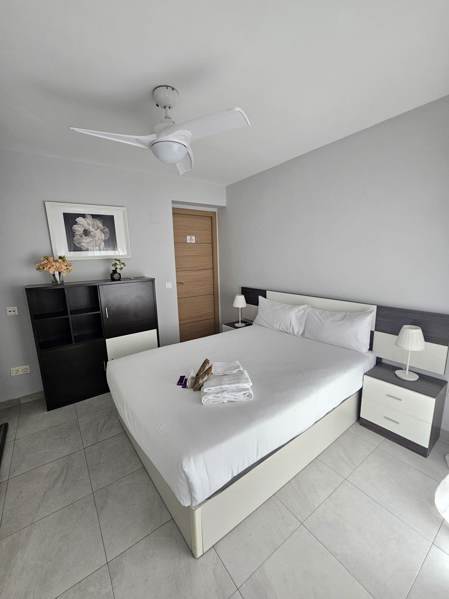Una habitación de hotel moderna con una cama grande, una mesita de noche, un armario y un ventilador de techo, además de un cuadro en la pared.