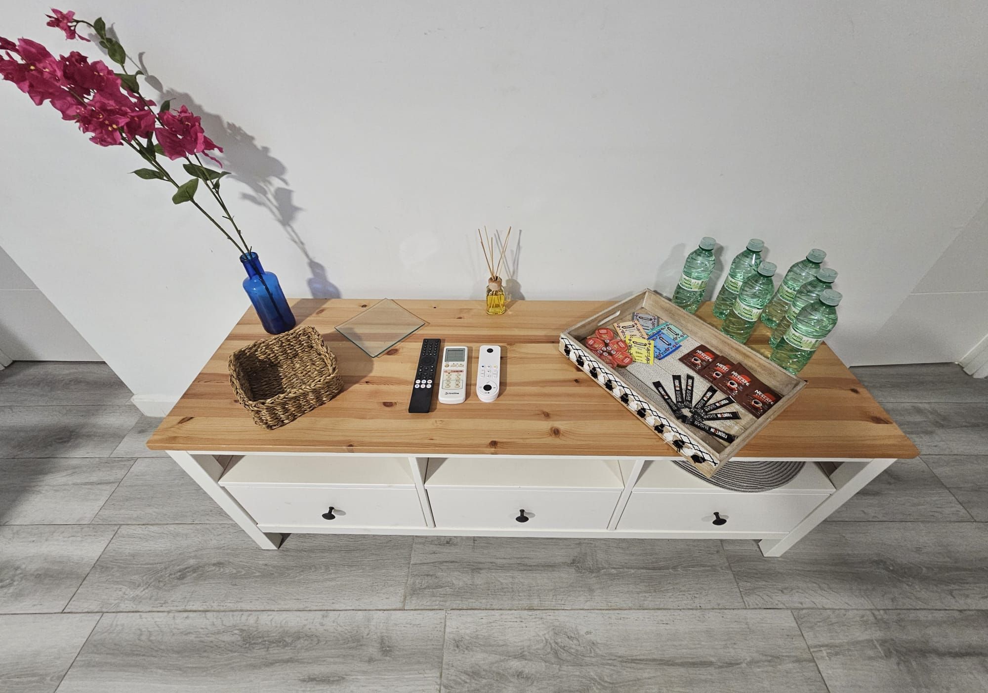 A wooden table with a vase, remote controls, a diffuser, and a tray of snacks and drinks.