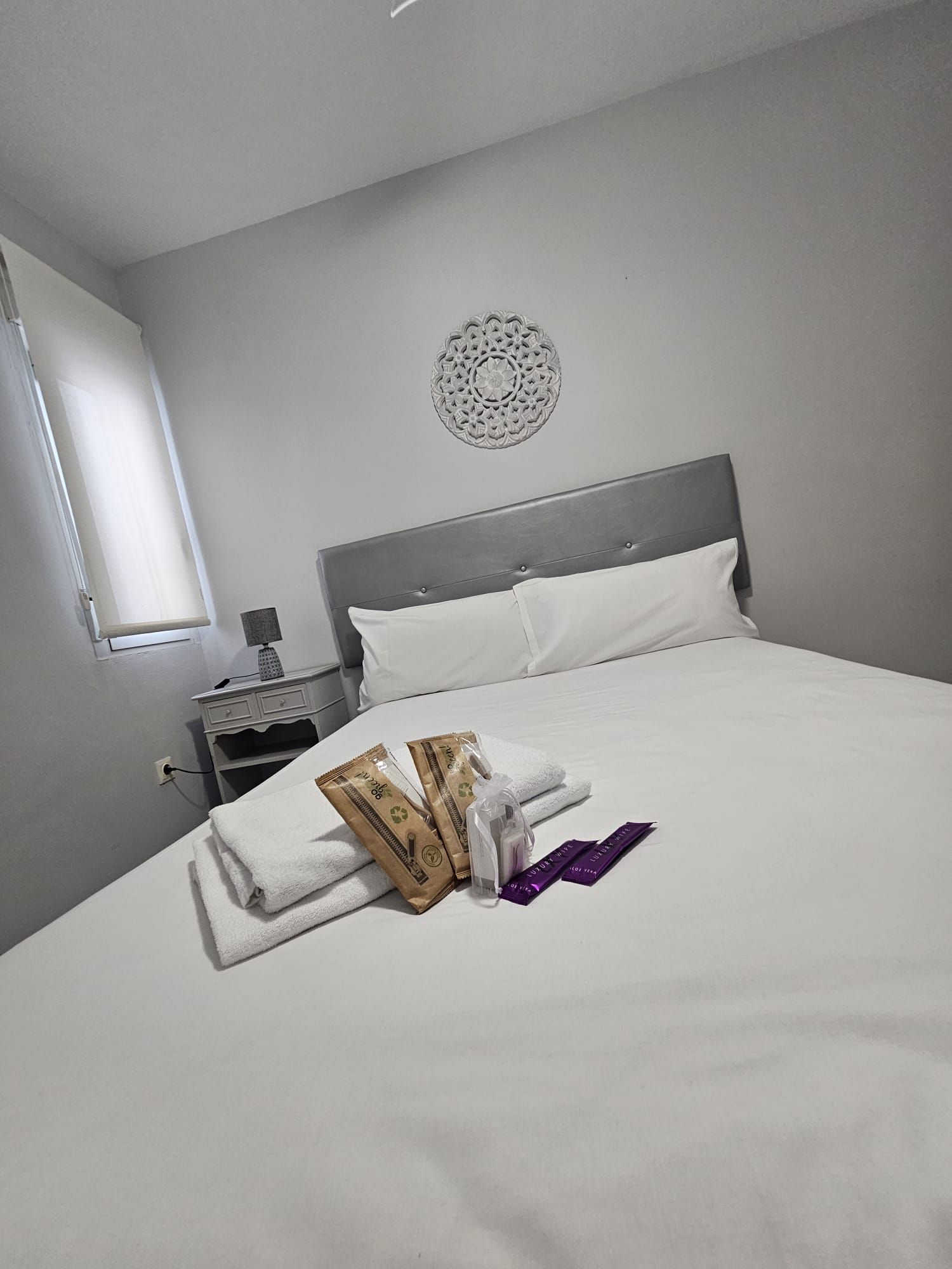 Una cama bien hecha con ropa blanca, un adorno en la pared y una mesita de noche con lámpara y un cajón pequeño.