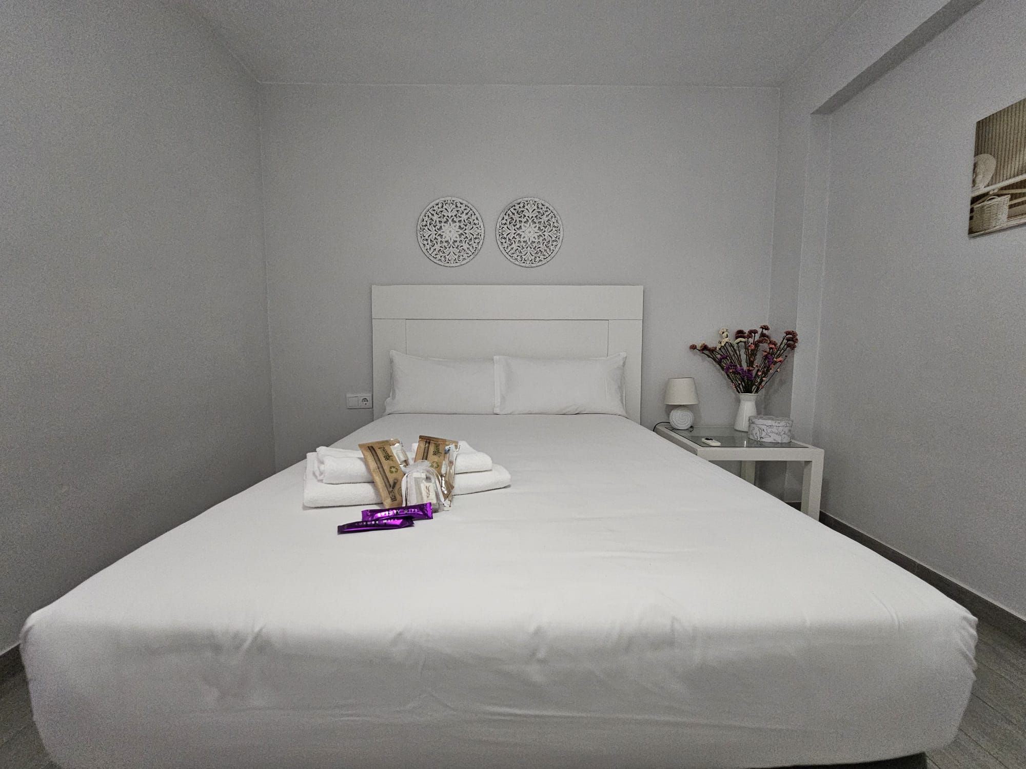 Una cama bien hecha con almohadas y una pequeña bolsa, una mesita de noche con una lámpara y un florero, y una pared con dos diseños circulares decorativos.