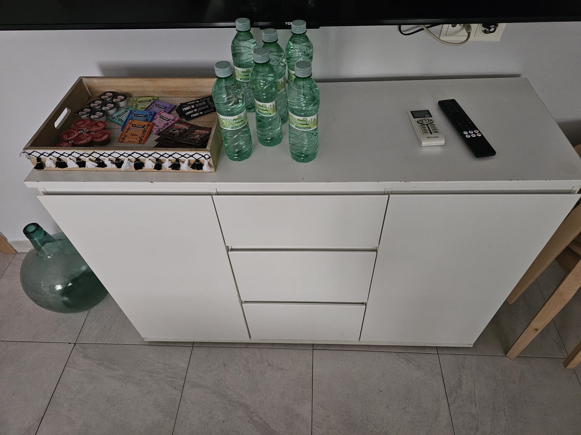 Een wit bureau met een lade, een afstandsbediening en verschillende groene flessen erop.