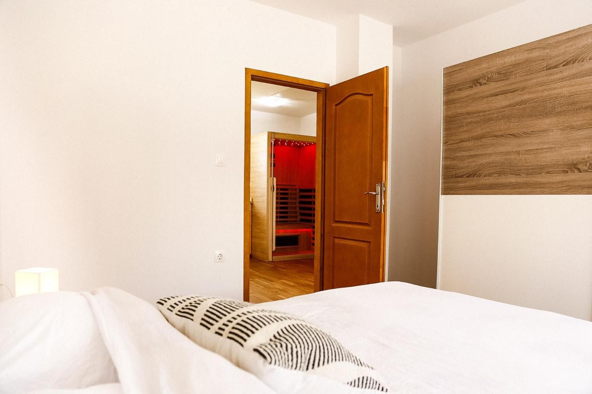 A bedroom with a bed, a wooden door, and a red-lit room in the background.