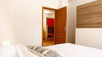 A bedroom with a bed, a wooden door, and a red-lit room in the background.