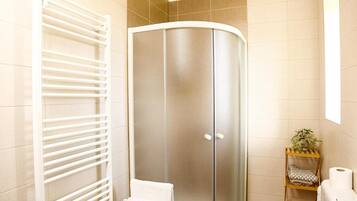 A bathroom with a glass shower enclosure, a white towel, a shelf with toiletries, and a white radiator.