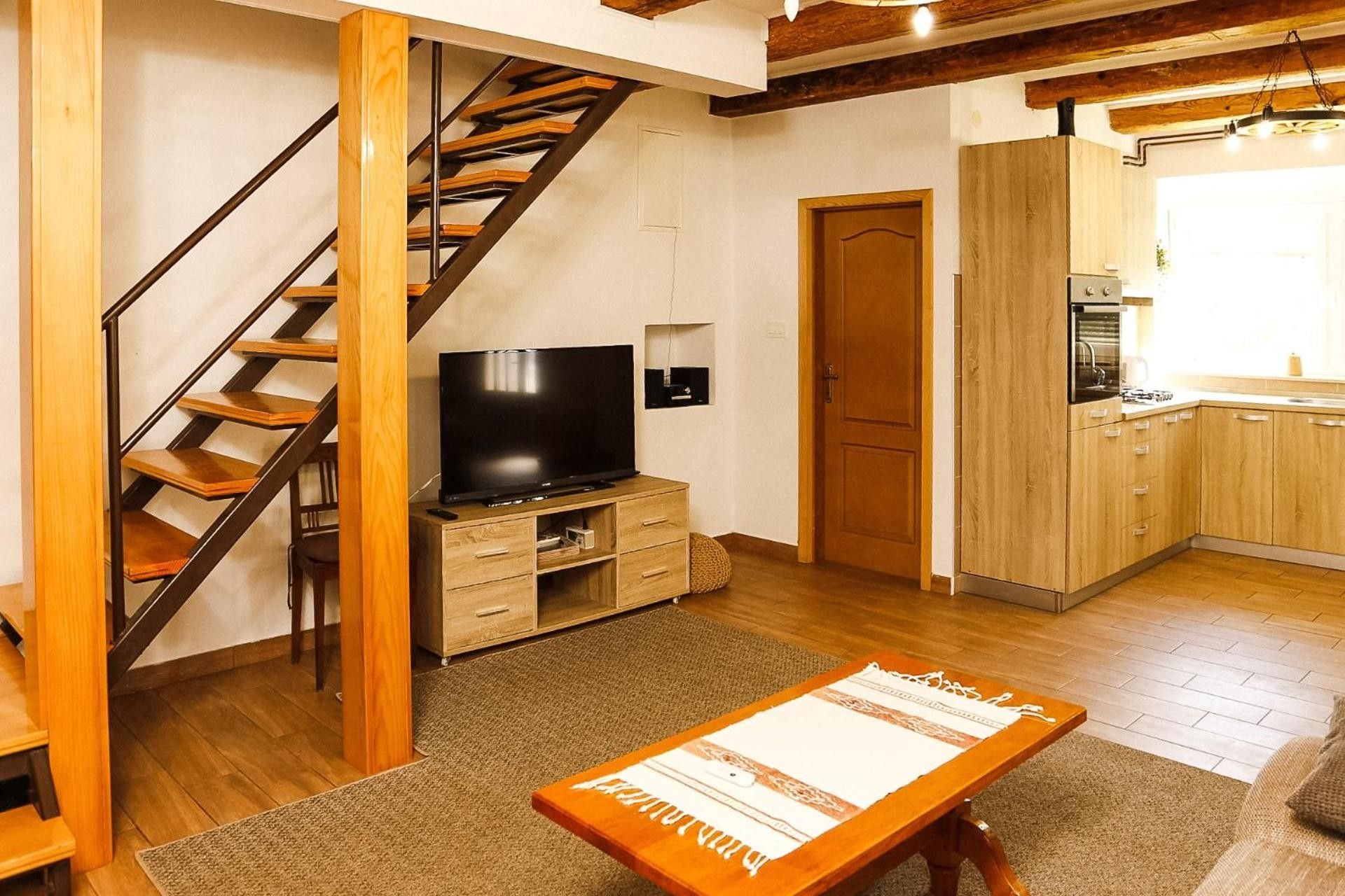 Ett modernt vardagsrum med en trätrappa, en platt-TV placerad på ett träskåp, ett köksområde med träskåp och ett matbord med en bordsduk.