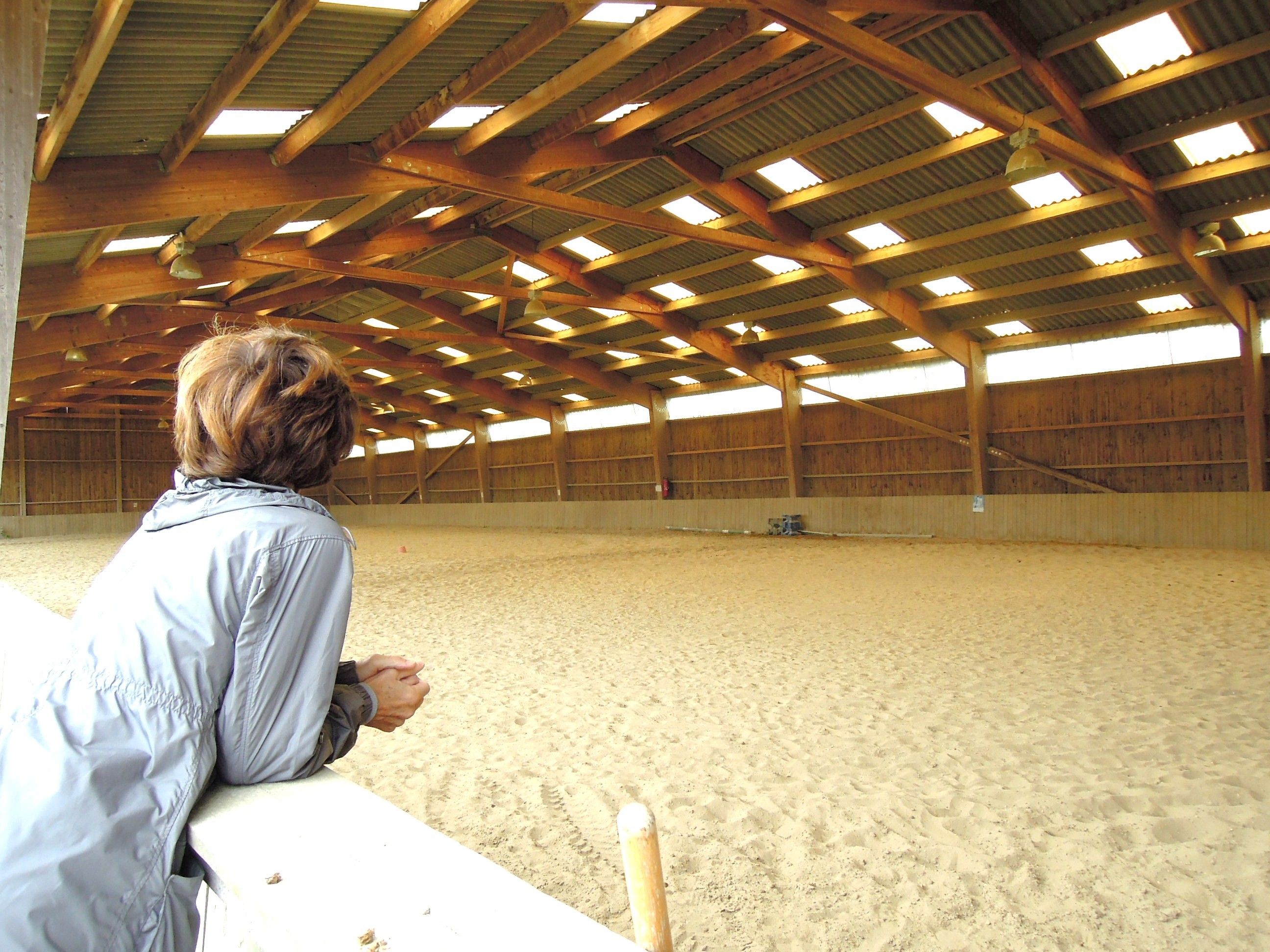 An indoor riding arena with a sandy floor, wooden beams, and a person observing from the side.