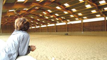 An indoor riding arena with a sandy floor, wooden beams, and a person observing from the side.