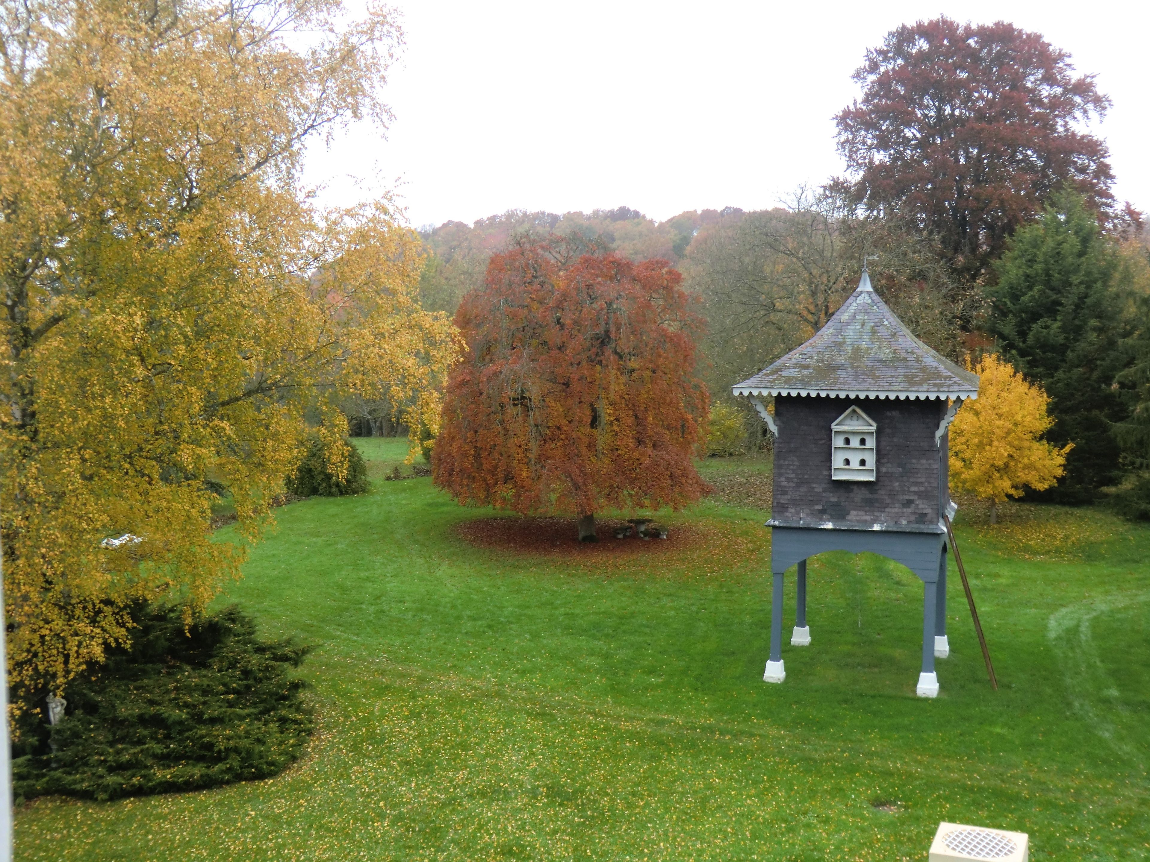 A small, standalone structure with a pointed roof, situated in a grassy area with trees displaying autumn foliage.
