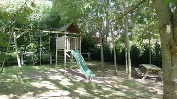 Une aire de jeux avec un toboggan et des balançoires, entourée d’arbres.
