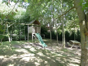 Un parque infantil con tobogán y columpios, rodeado de árboles.