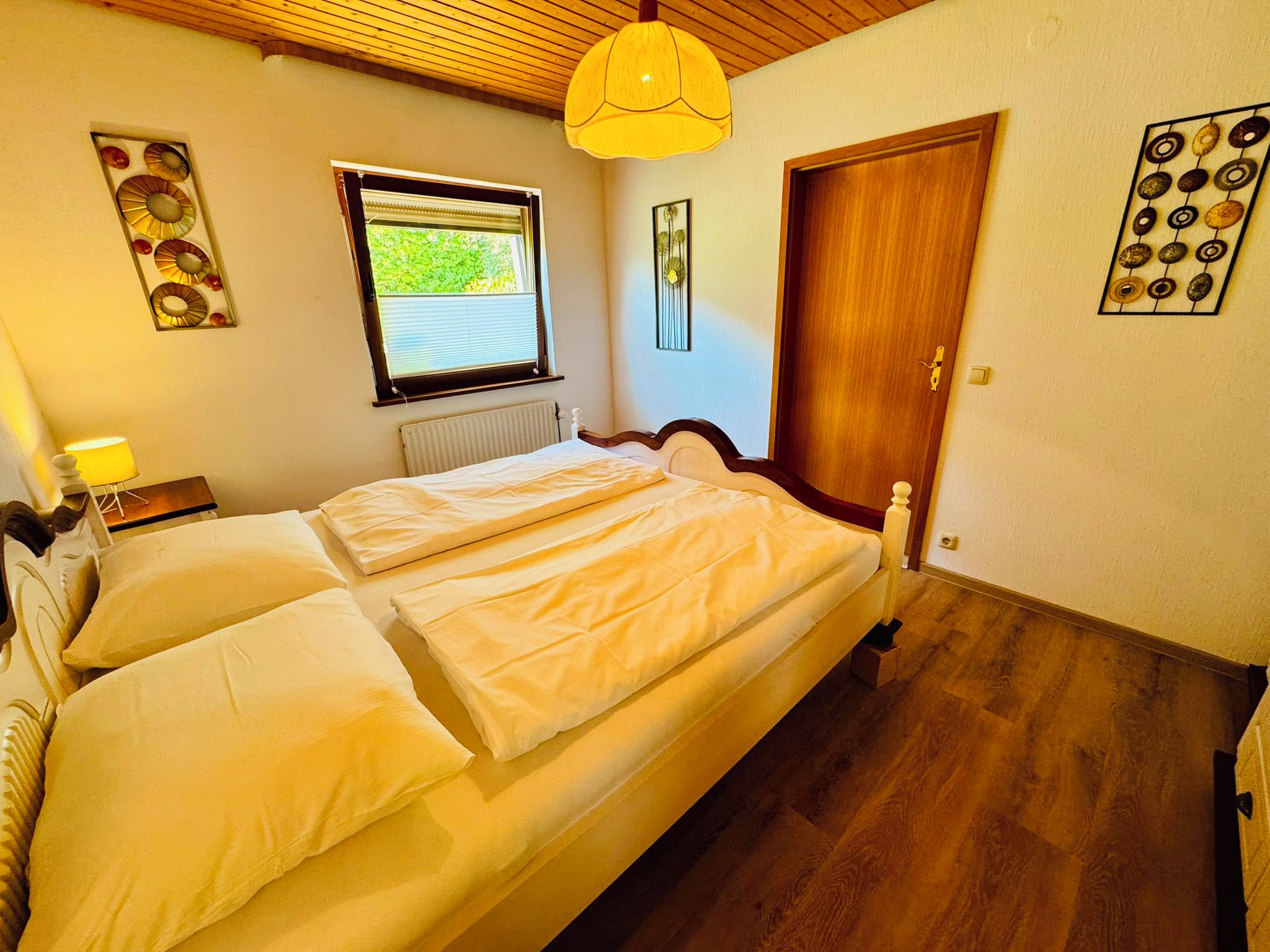 Een slaapkamer met een tweepersoonsbed, een raam, een radiator, een houten deur en een wandlamp.