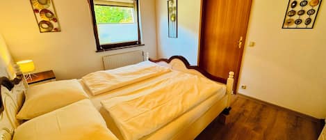 A bedroom with a double bed, a window, a radiator, a wooden door, and a wall-mounted light fixture.