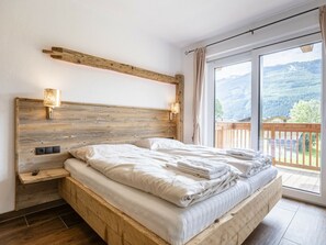 Un dormitorio con cabecera de madera, dos lámparas de noche, un ventanal con vistas a las montañas y un balcón.