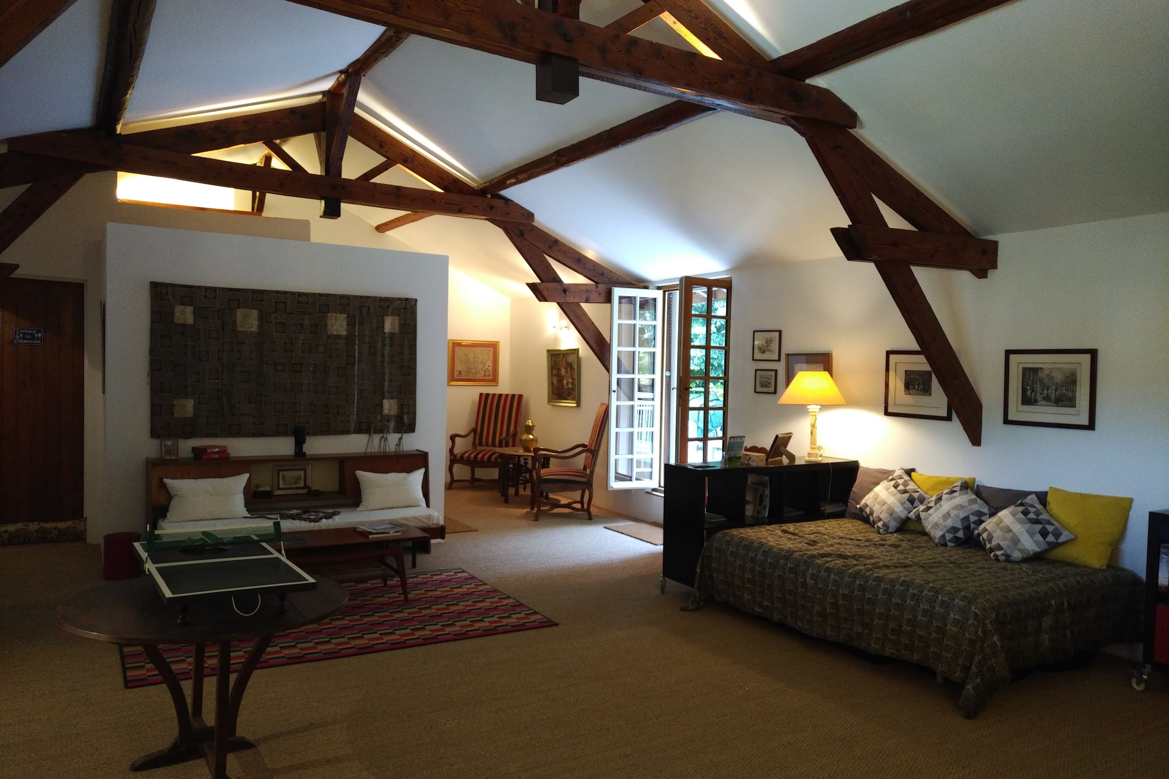 A cozy living room with a high ceiling, exposed wooden beams, a sofa, a coffee table, a dining area with chairs, and a bed with patterned bedding.