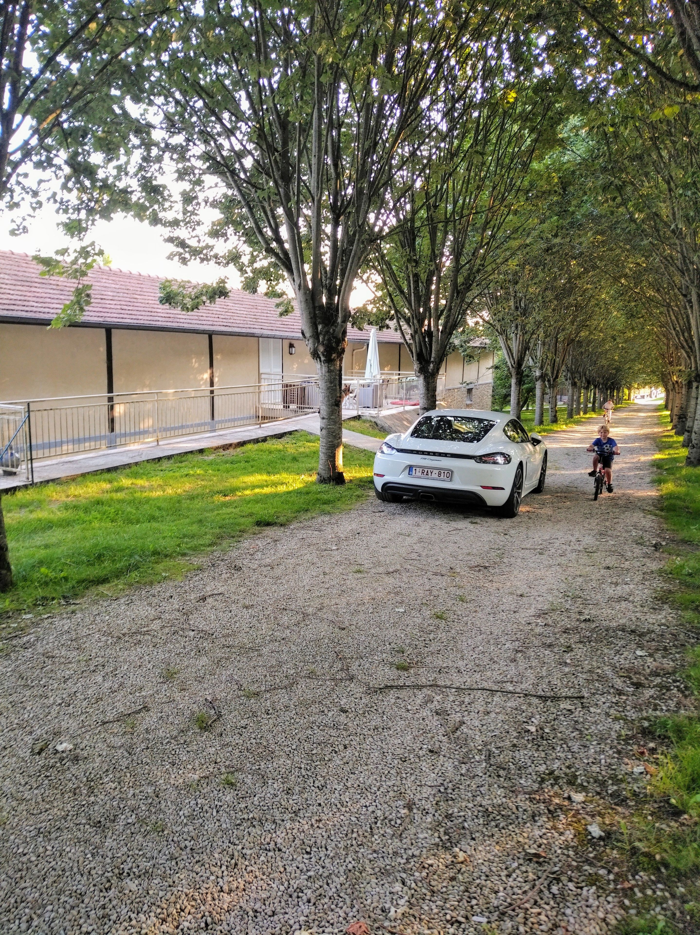 Un camino de grava con un coche blanco estacionado al costado, árboles bordeando el sendero y un edificio con techo rojo al fondo.