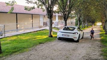 Un camino de grava con un coche blanco estacionado al costado, árboles bordeando el sendero y un edificio con techo rojo al fondo.