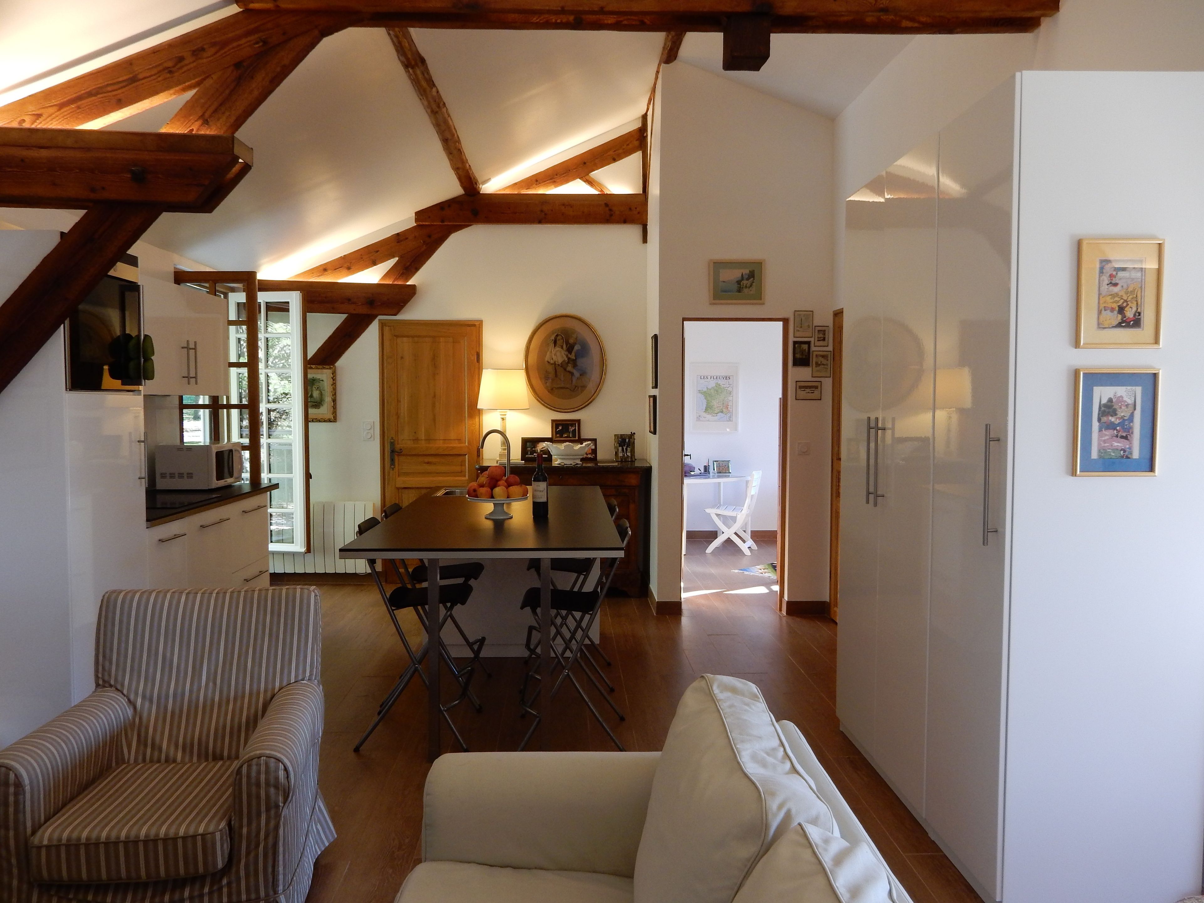 Een ruime woonkamer met een hoge plafond en zichtbare houten balken, een eettafel met stoelen, een bank en een keuken met een koelkast.