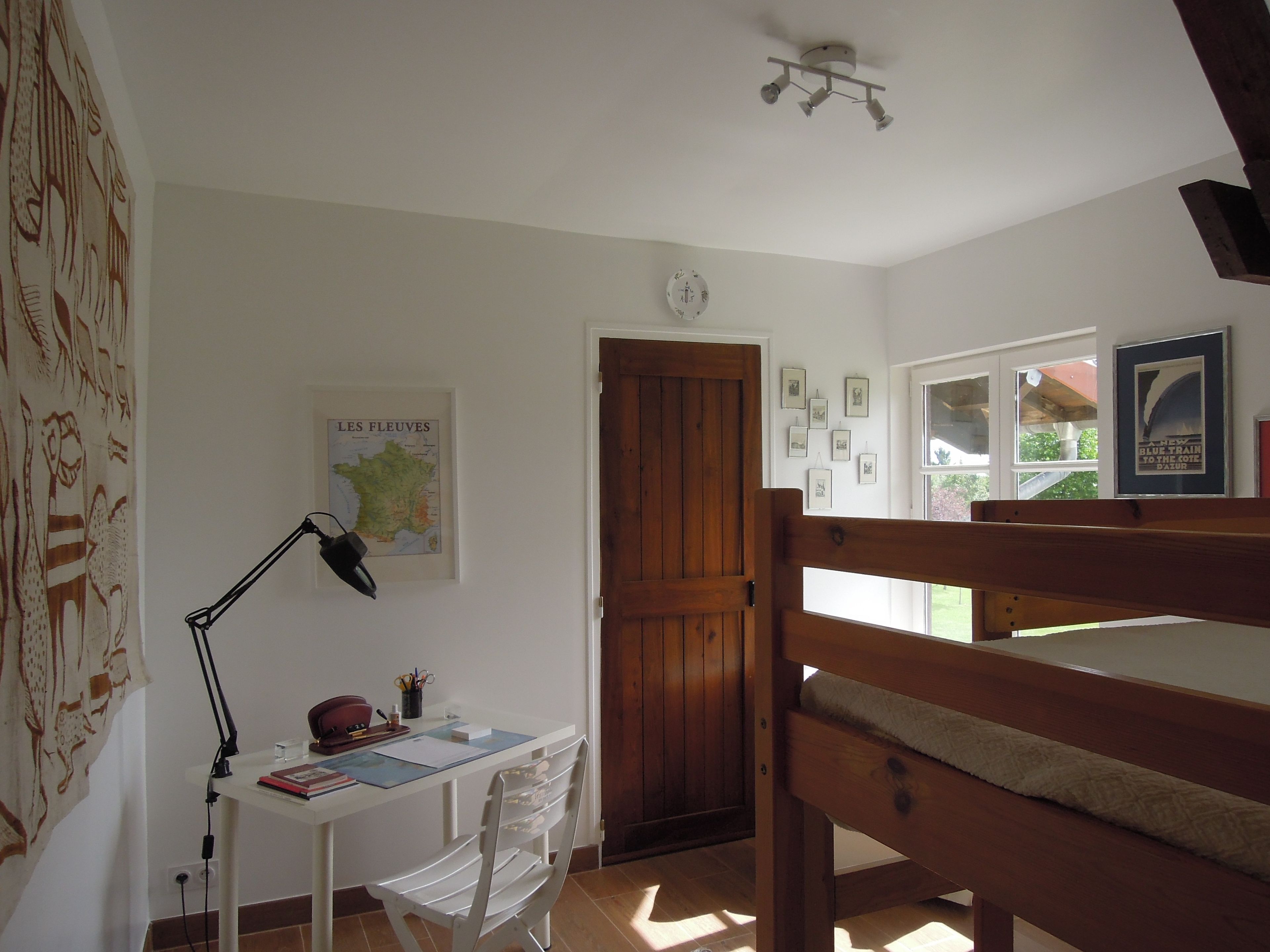 Una habitación con una cama de madera, un escritorio con una lámpara, una silla, un mapa en la pared y una puerta que da a otra habitación.