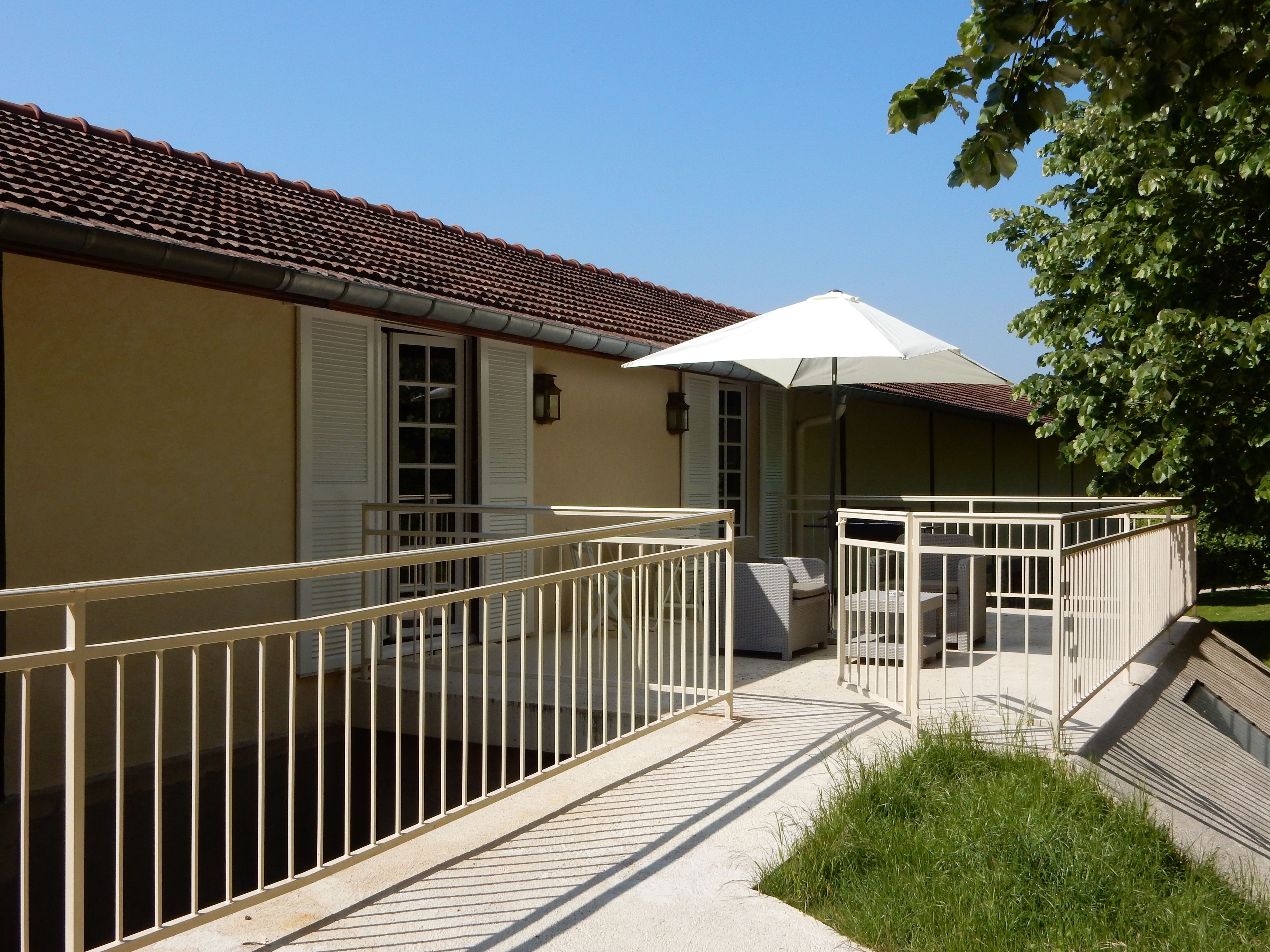 Un bâtiment avec un toit recouvert de tuiles, un balcon doté d’une balustrade blanche et un espace de repos couvert avec un parasol blanc.