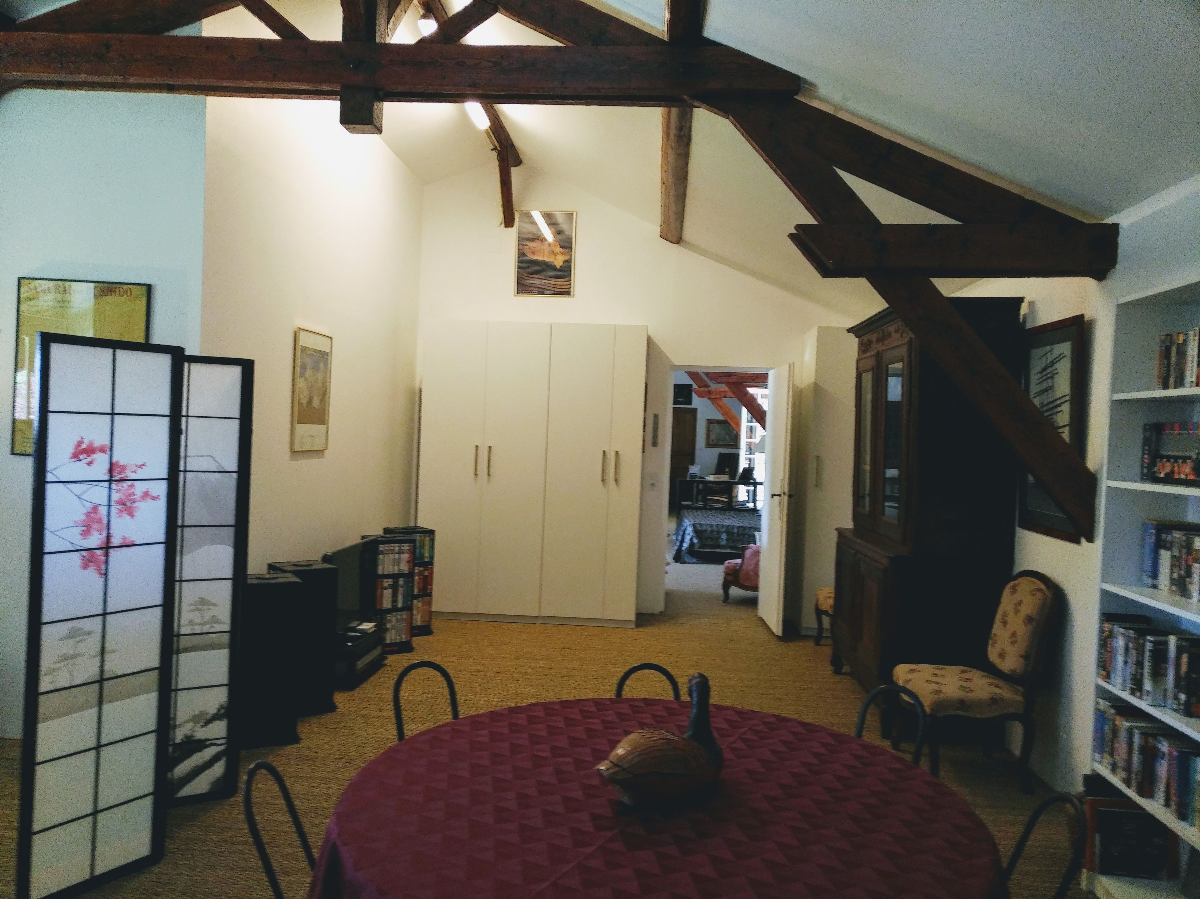 Una habitación con techo alto, vigas de madera, una mesa redonda con mantel rojo, una estantería y un biombó tradicional japonés.