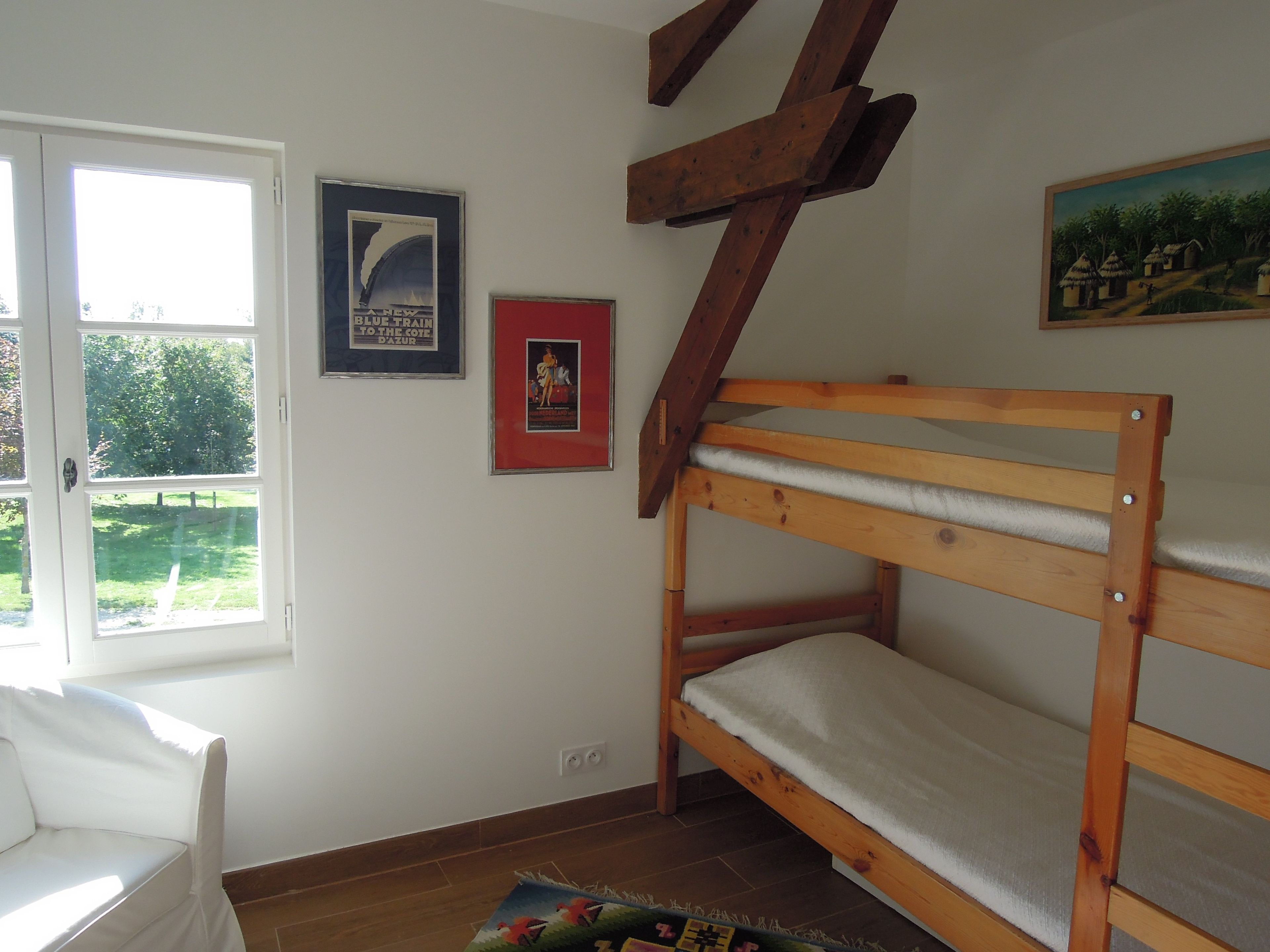A room with a bunk bed, a white sofa, a window with a view of greenery, and two framed pictures on the wall.