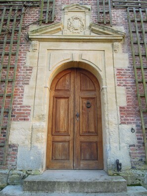 Una puerta doble de madera con un arco de piedra, integrada en un edificio de ladrillo con un escudo decorativo en la parte superior.