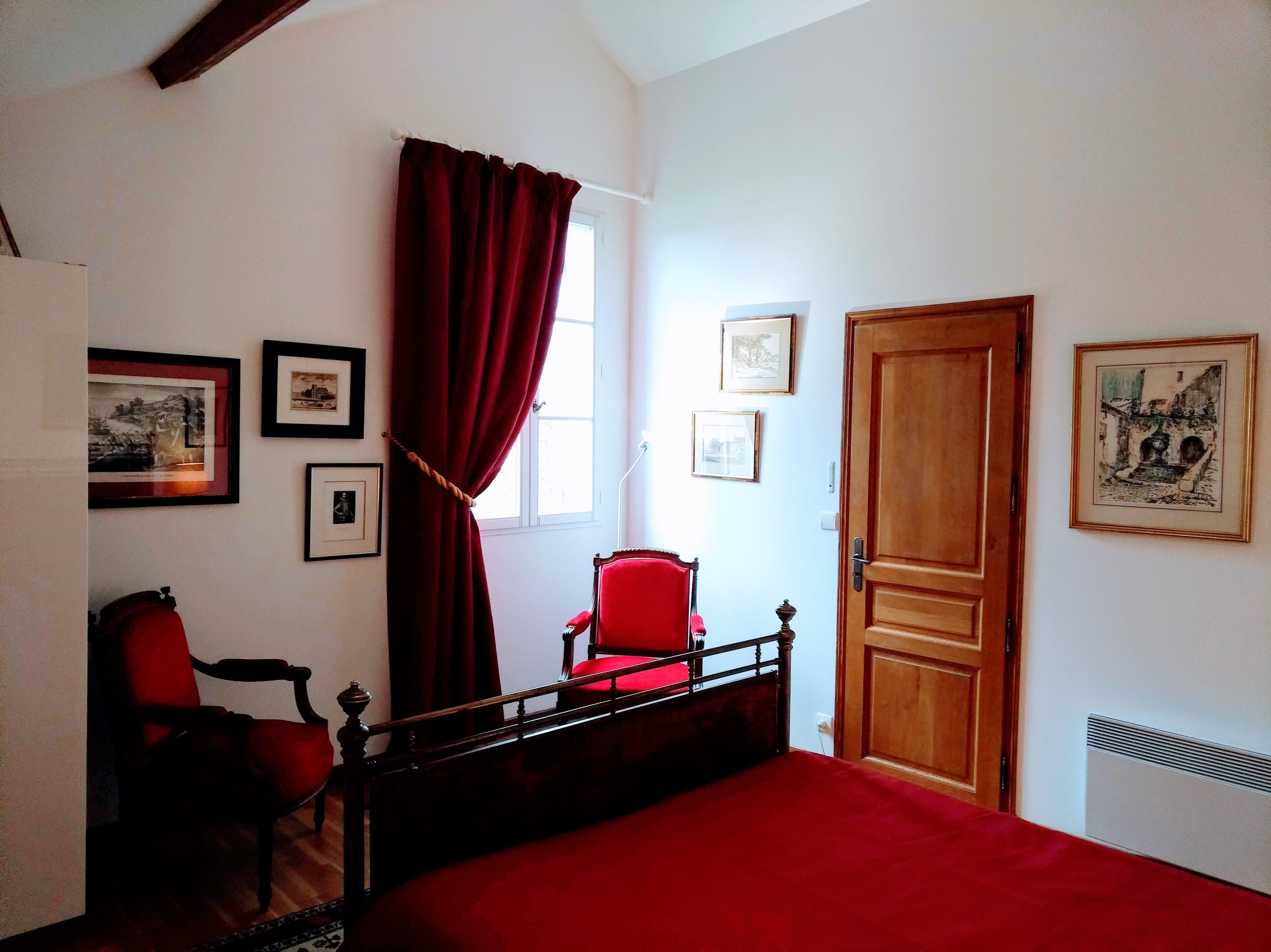 A bedroom with a bed, a red chair, a wooden door, and framed pictures on the walls.