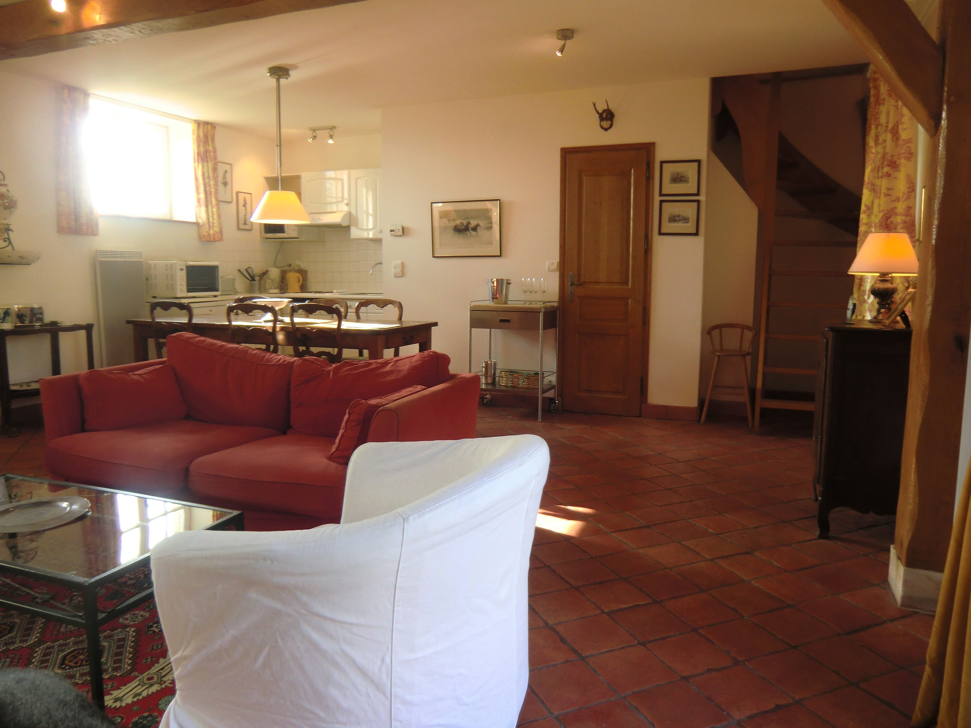 Una sala de estar con un sofá rojo, un sillón blanco, una mesa de centro de vidrio y una cocina al fondo.