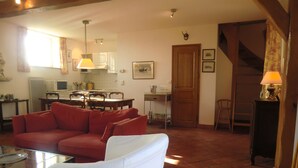 Un salon avec un canapé rouge, un fauteuil blanc, une table basse en verre et, au fond, un coin cuisine.