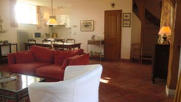 Una sala de estar con un sofá rojo, un sillón blanco, una mesa de centro de vidrio y una cocina al fondo.
