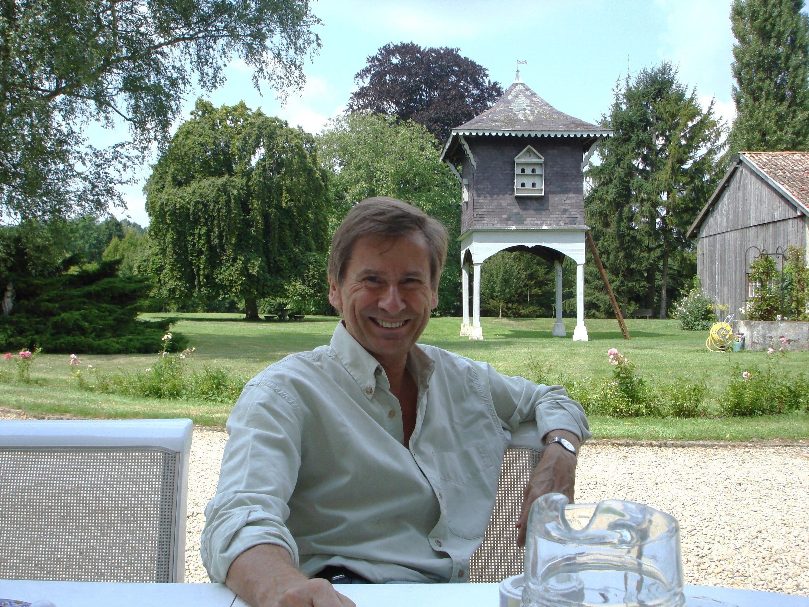 A man seated at an outdoor table with a clear view of a gazebo and a barn in the background.