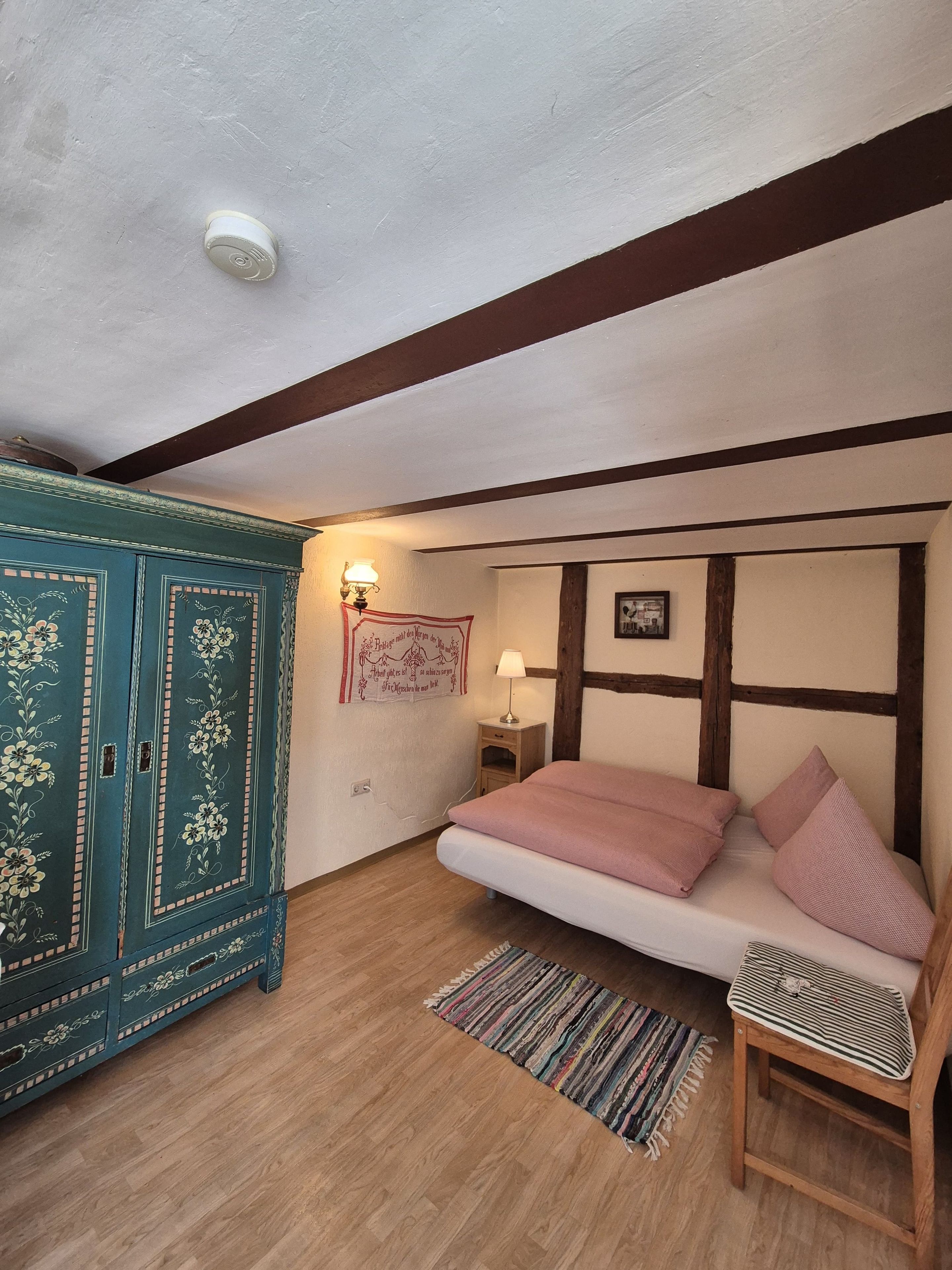 Une chambre à coucher comprenant un lit, une chaise en bois, une petite table avec une lampe, un meuble décoratif et un mur orné d’un tableau encadré.